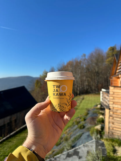 Kubek papierowy z żółtym nadrukiem „TW JA O KAWA” trzymany w dłoni